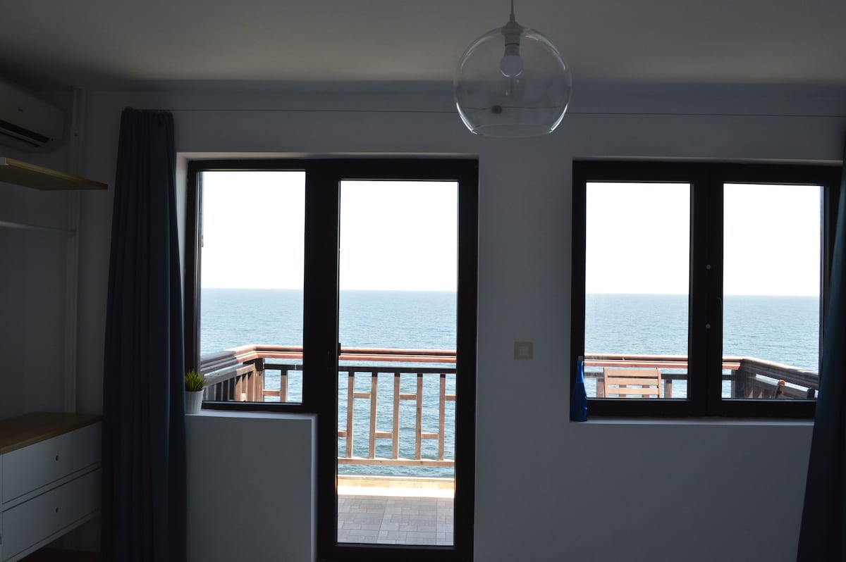 Two large windows frame a stunning view of the sea, allowing natural light to fill the room. The wooden balcony railing can be seen beyond, creating a seamless connection to the outdoor space.