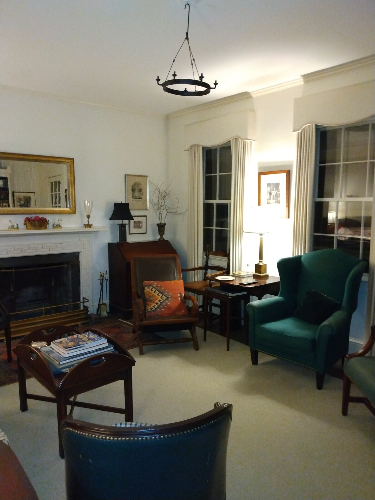 A comfortable living room is presented with an inviting fireplace, surrounded by windows that allow natural light to fill the space. Green upholstered chairs, a wooden side table, and decorative accents create a cozy atmosphere for relaxation.