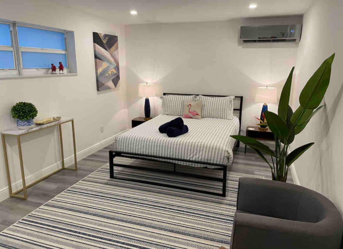 A serene bedroom features a spacious bed adorned with striped bedding and a navy throw. Soft lighting is provided by bedside lamps. A large green plant adds a touch of nature, while a neatly arranged rug spans the floor, enhancing the room's contemporary design.