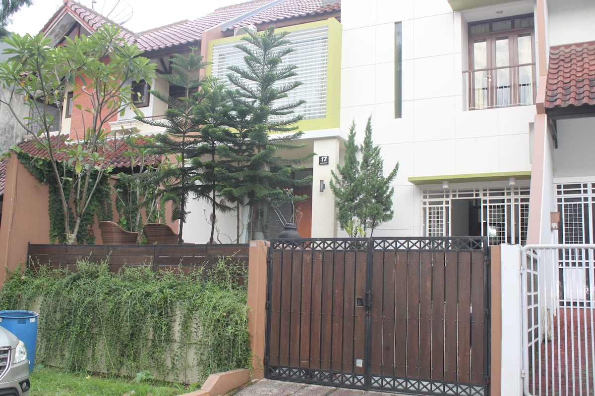 The exterior of the holiday home features a modern design, with a mix of greenery and a brown wooden gate. Potted plants and a small sitting area are visible on the porch, while a mix of tile and brick accents adorn the facade.