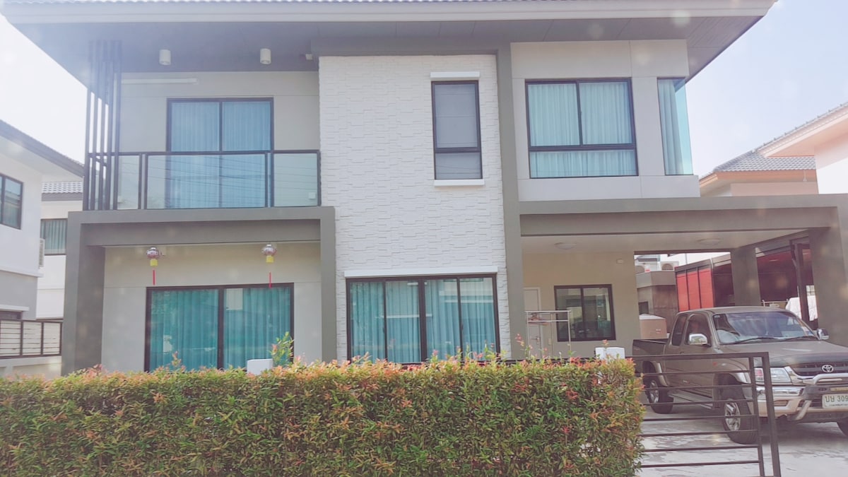 A two-story building features a modern design with large glass windows and a textured white facade. A lush green hedge lines the front, offering a sense of privacy. The covered parking area accommodates a vehicle, contributing to the home's functionality.