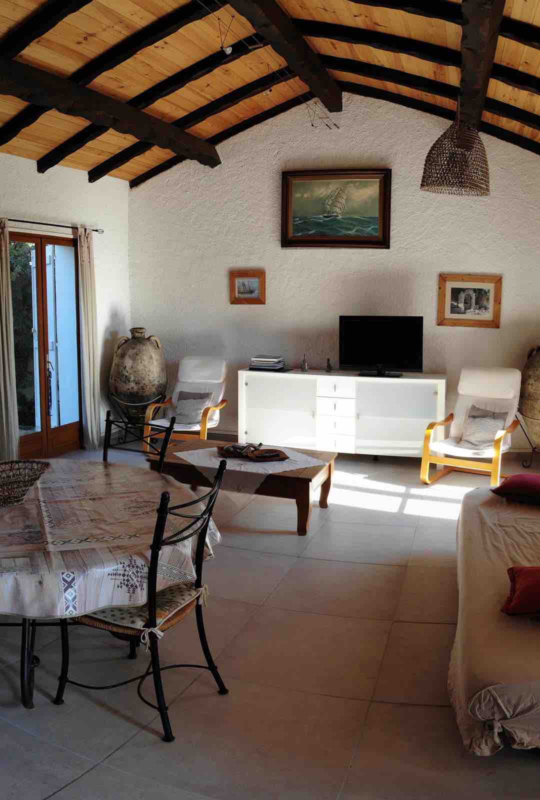 A spacious living area features high, wooden-beamed ceilings and natural light from multiple windows. A round dining table with chairs is positioned next to a cozy seating area that includes two armchairs and a couch. A television sits atop a white cabinet, and decorative elements enhance the calm environment.