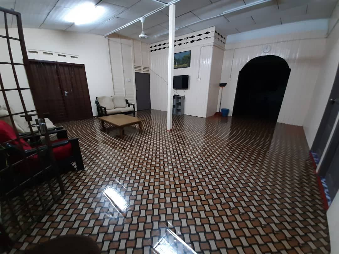 A spacious living area features a patterned tiled floor and is furnished with wooden chairs and a low table. A wall-mounted television is positioned near a door, while light from overhead fixtures illuminates the room.