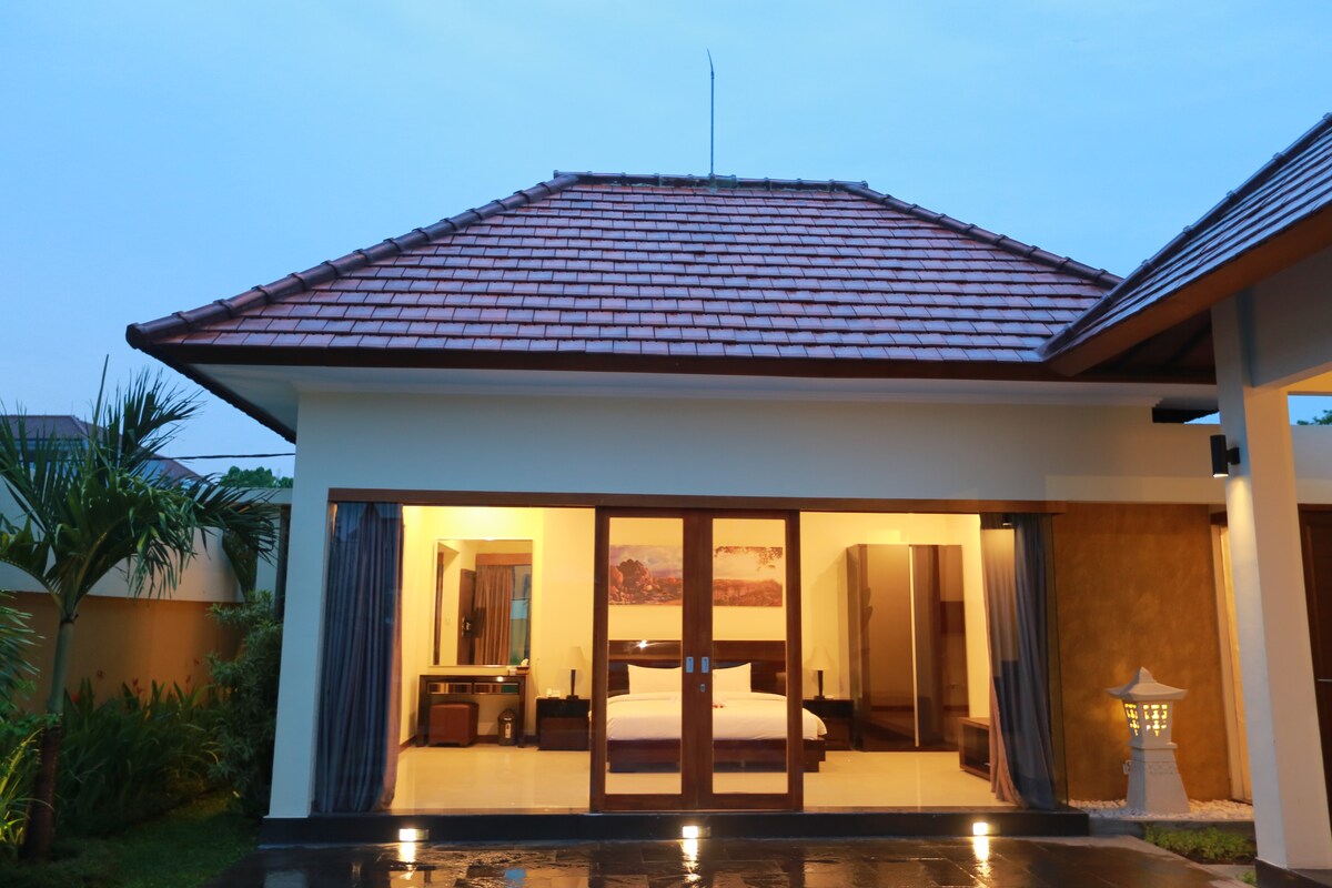 The exterior of a villa is shown, featuring a traditional roof design with terracotta tiles. Large glass doors reveal a spacious interior, with soft lighting highlighting the furnishings inside. Surrounding greenery enhances the serene privacy of the property.