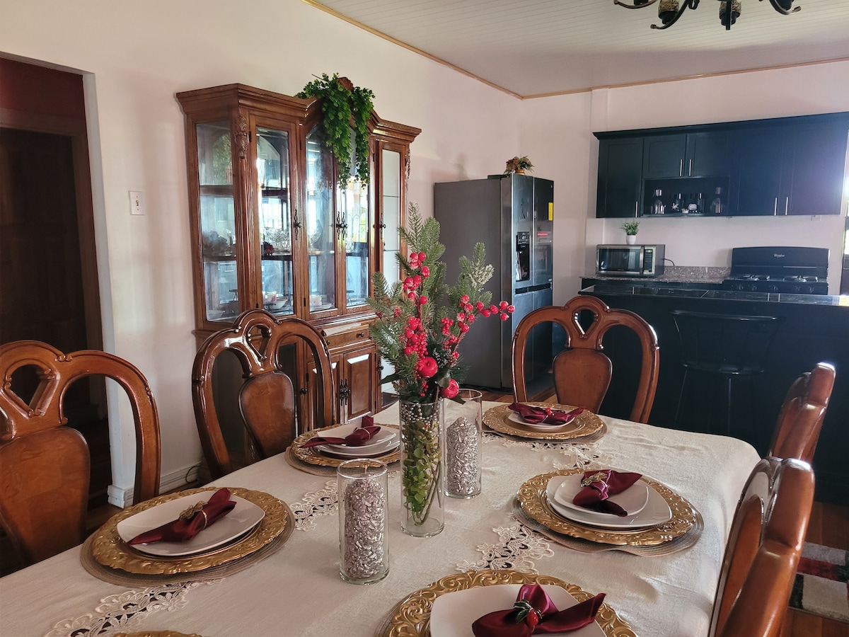 A dining area is highlighted with an elegant table set for six, featuring decorative plates and napkins. A glass display cabinet and modern kitchen appliances are visible in the backdrop, with greenery adorning the space, contributing to a refined ambiance.