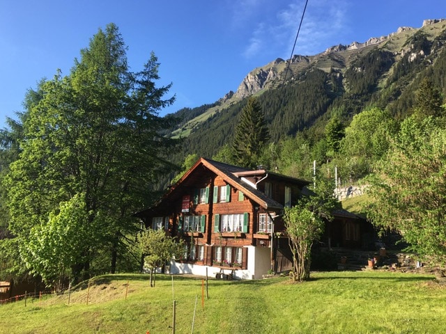 A charming Swiss chalet is set against a backdrop of mountains and blue skies. The wooden structure features multiple floors with green shutters and a welcoming appearance. Surrounding trees and landscaped grounds enhance the serene environment.