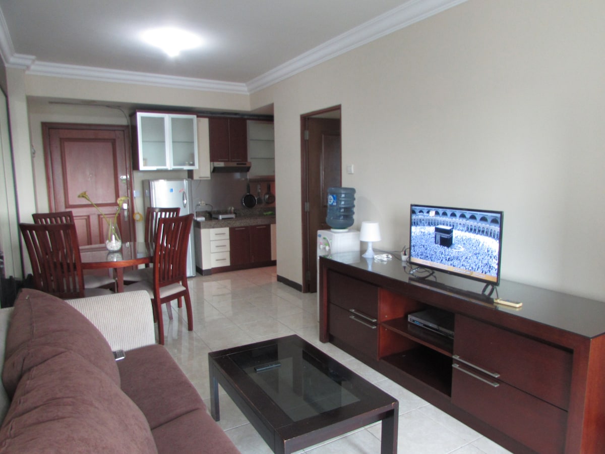 The living area features a comfortable brown sofa and a glass coffee table. A wooden cabinet holds a television, which displays a scene of a large gathering. In the background, a dining table with four chairs and a kitchen area are visible.