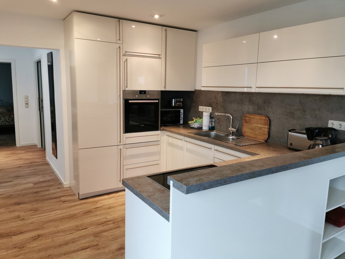 A modern kitchen features sleek white cabinetry and a gray countertop. Stainless steel appliances, including an oven, microwave, and coffee maker, are visible. Bright lighting enhances the space, which offers a clear view into the adjoining living area through an open layout.