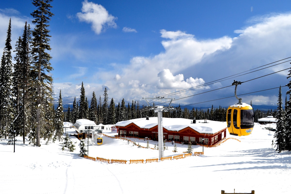 A bright, snowy landscape features a yellow gondola gliding above a cozy lodge with red accents. Towering evergreens frame the scene against a backdrop of expansive blue skies and fluffy white clouds, creating a serene atmosphere in a winter wonderland.