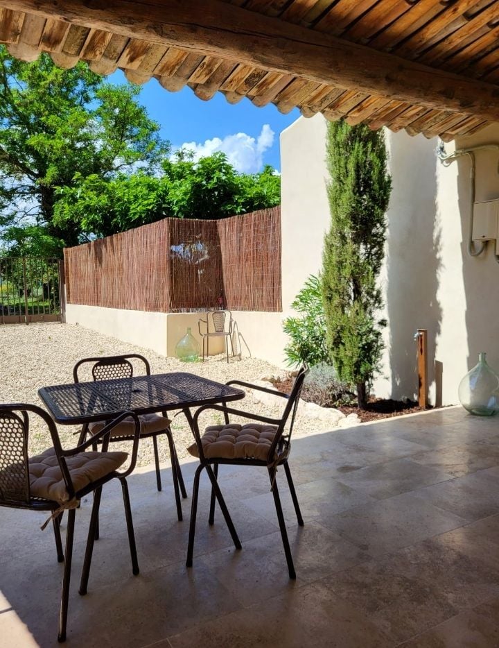An outdoor terrace is furnished with a small table and two chairs, situated under a wooden canopy. A gravel area surrounds the terrace, complemented by a tall shrub and a bamboo privacy screen in the background, with green trees visible under a clear blue sky.