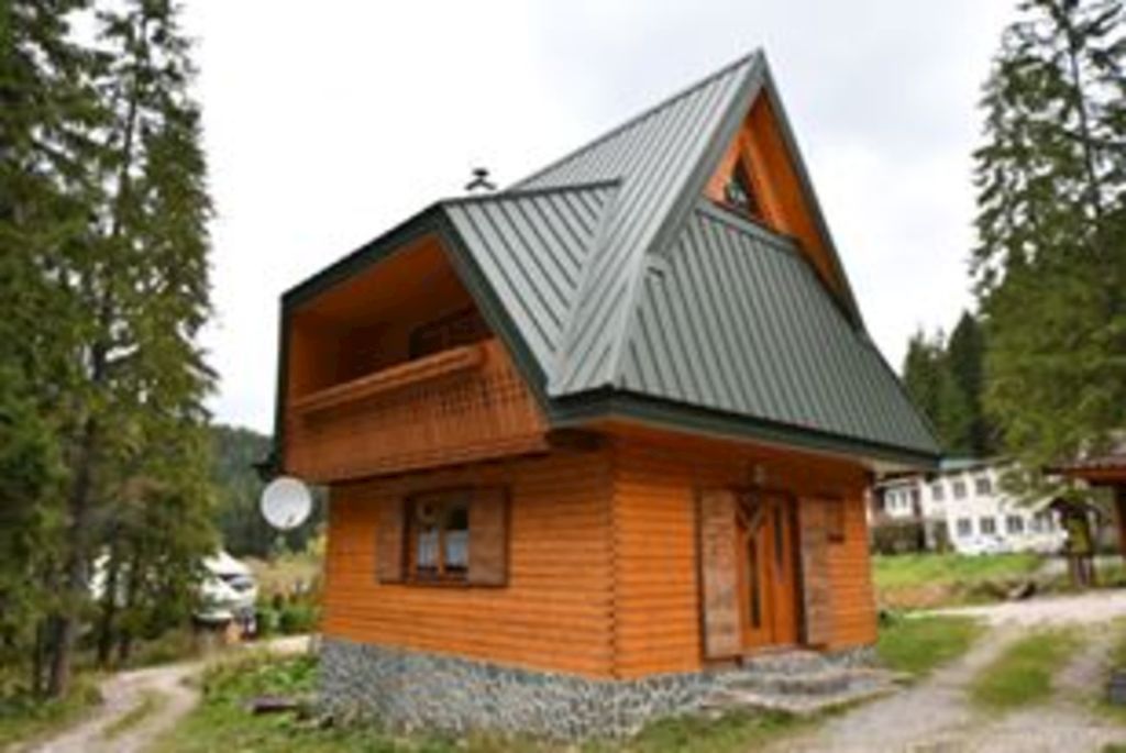 A charming wooden chalet with a steeply pitched metal roof is surrounded by greenery. A spacious balcony is featured on the upper level, and a stone foundation complements the wooden walls. Nearby trees add to the serene environment of the accommodation.