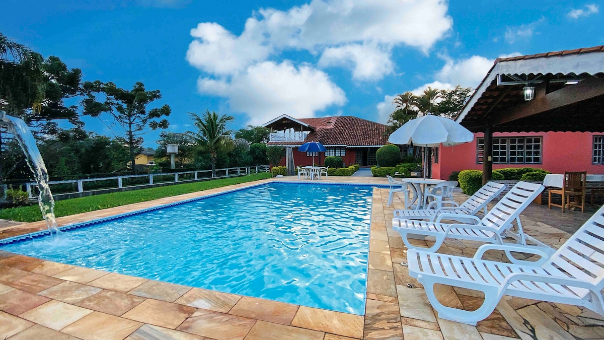 A clear blue swimming pool is surrounded by lounge chairs and a shaded umbrella. Lush greenery and trees frame the scene, while a charming house sits in the background. Sunlight reflects on the water, creating a serene outdoor space for relaxation.
