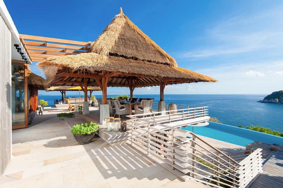 An outdoor area features a thatched roof structure with a wooden framework, providing shade over a dining table surrounded by woven chairs. The infinity pool reflects the blue sky and ocean, while tropical plants add greenery to the serene atmosphere.