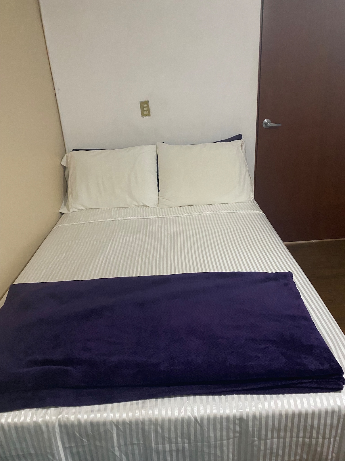 A neat sleeping area is presented, featuring a spacious matrimonial bed and an individual bed, both dressed in crisp white bedding. A plush purple blanket is neatly arranged at the foot of the beds. A wooden door is visible in the background.