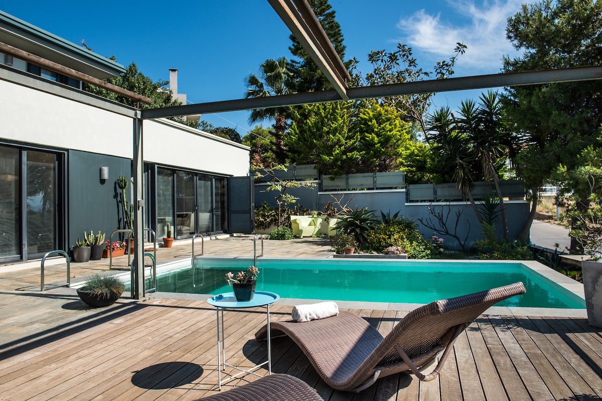 A private outdoor area features a swimming pool surrounded by lush greenery. Wooden loungers provide seating alongside a small table. Large glass doors invite natural light, creating an airy and harmonious atmosphere with ample space for relaxation and enjoyment.