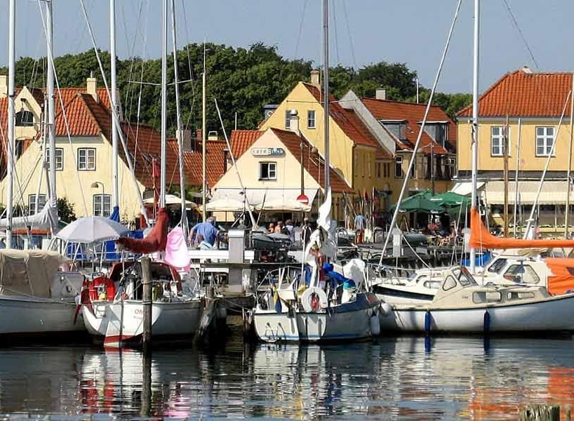 The harbor scene captures a number of sailboats docked in calm waters, reflecting the charming yellow buildings with red roofs lining the shore. The atmosphere is enhanced by the presence of people enjoying leisure time, with greenery visible in the background.