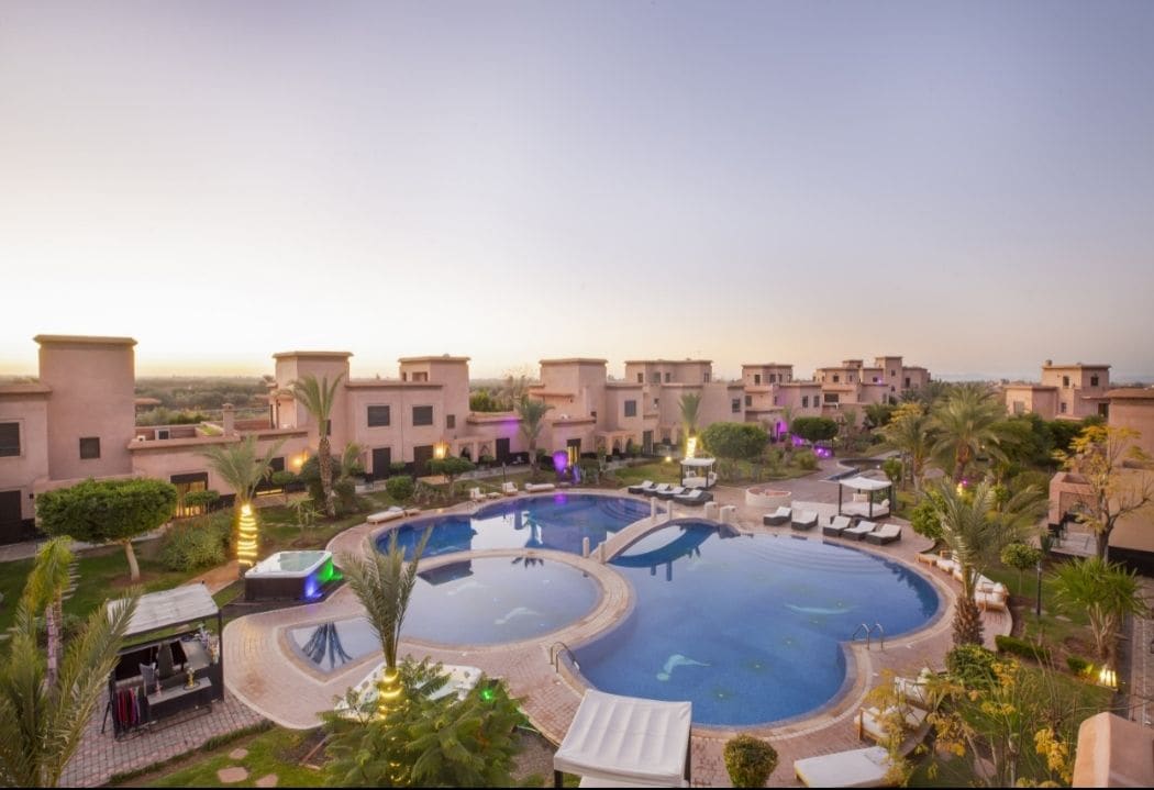The image captures a spacious outdoor area featuring a large swimming pool surrounded by tall palm trees and sun loungers. The tranquil water reflects the evening sky, while nearby villas are visible in the background, creating a peaceful resort atmosphere.