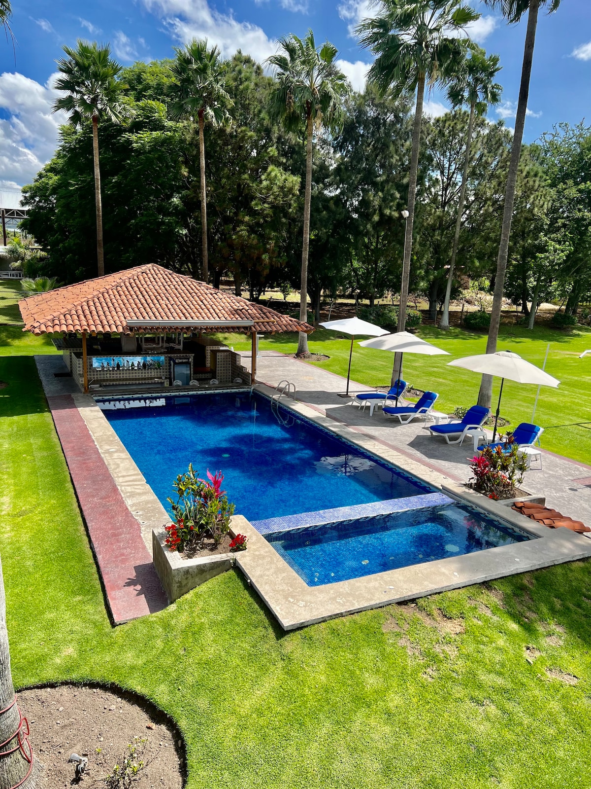 A private outdoor area features a heated swimming pool surrounded by lush greenery. Two lounge chairs and umbrellas are positioned near the water, while a shaded bar with seating adds to the leisure space. Colorful flower beds enhance the serene environment.