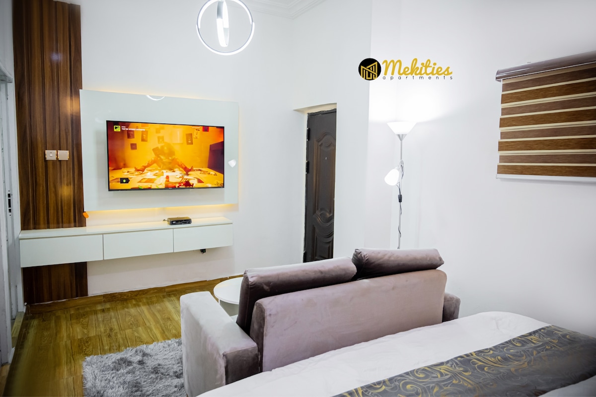 A modern living area is shown, featuring a wall-mounted flat-screen television with a vibrant display. A cozy gray sofa is positioned in front, complemented by natural wood flooring. Soft lighting fixtures provide illumination, enhancing the overall ambiance of the space.