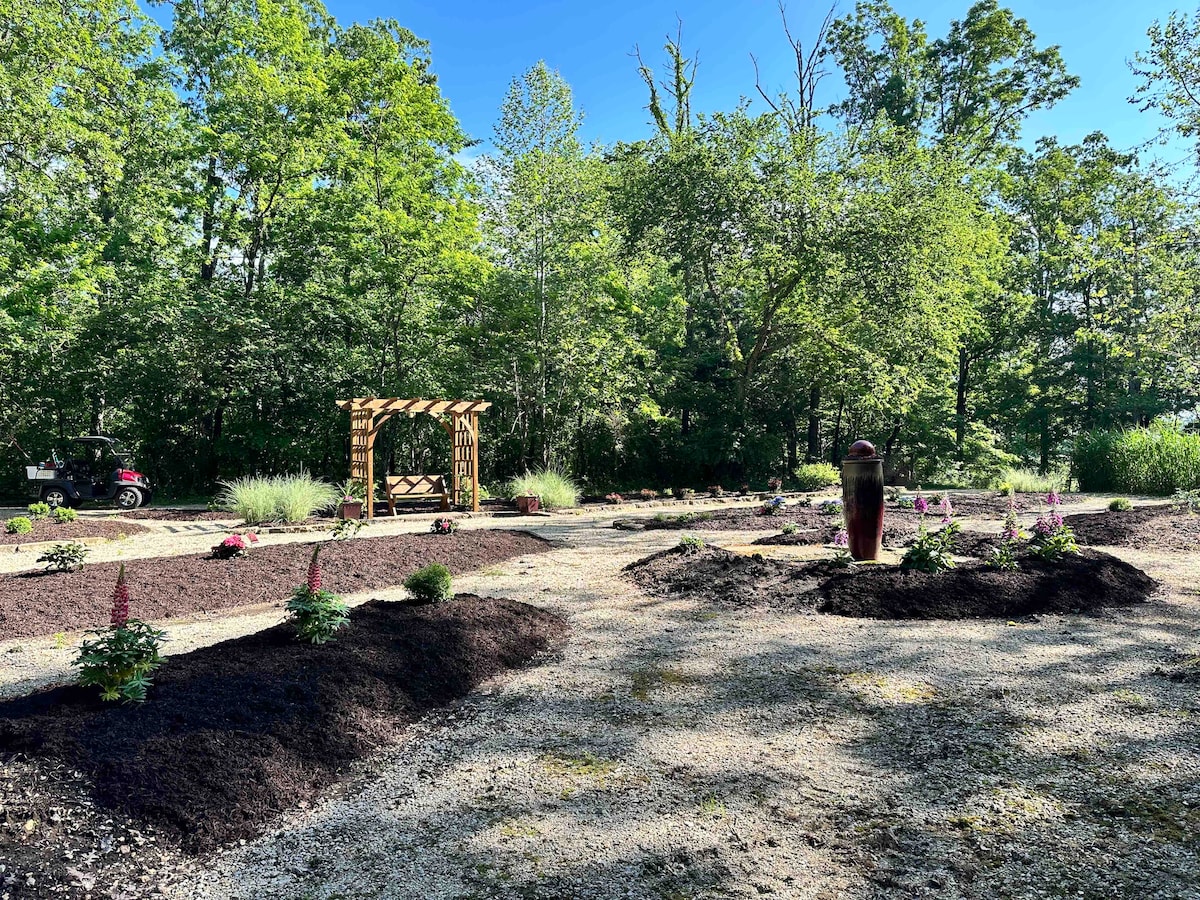 A landscaped area features gravel paths surrounded by lush greenery, including trees and flowering plants. An arbor stands in the distance, providing a focal point. Decorative elements such as a tall sculpture add interest to the inviting outdoor space.