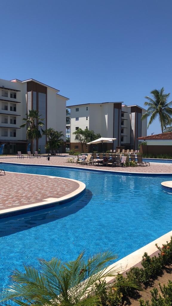 A spacious pool area is highlighted by a large, inviting blue pool surrounded by lounge chairs and umbrellas. The backdrop features contemporary buildings and palm trees under a clear blue sky, enhancing the serene environment ideal for relaxation.