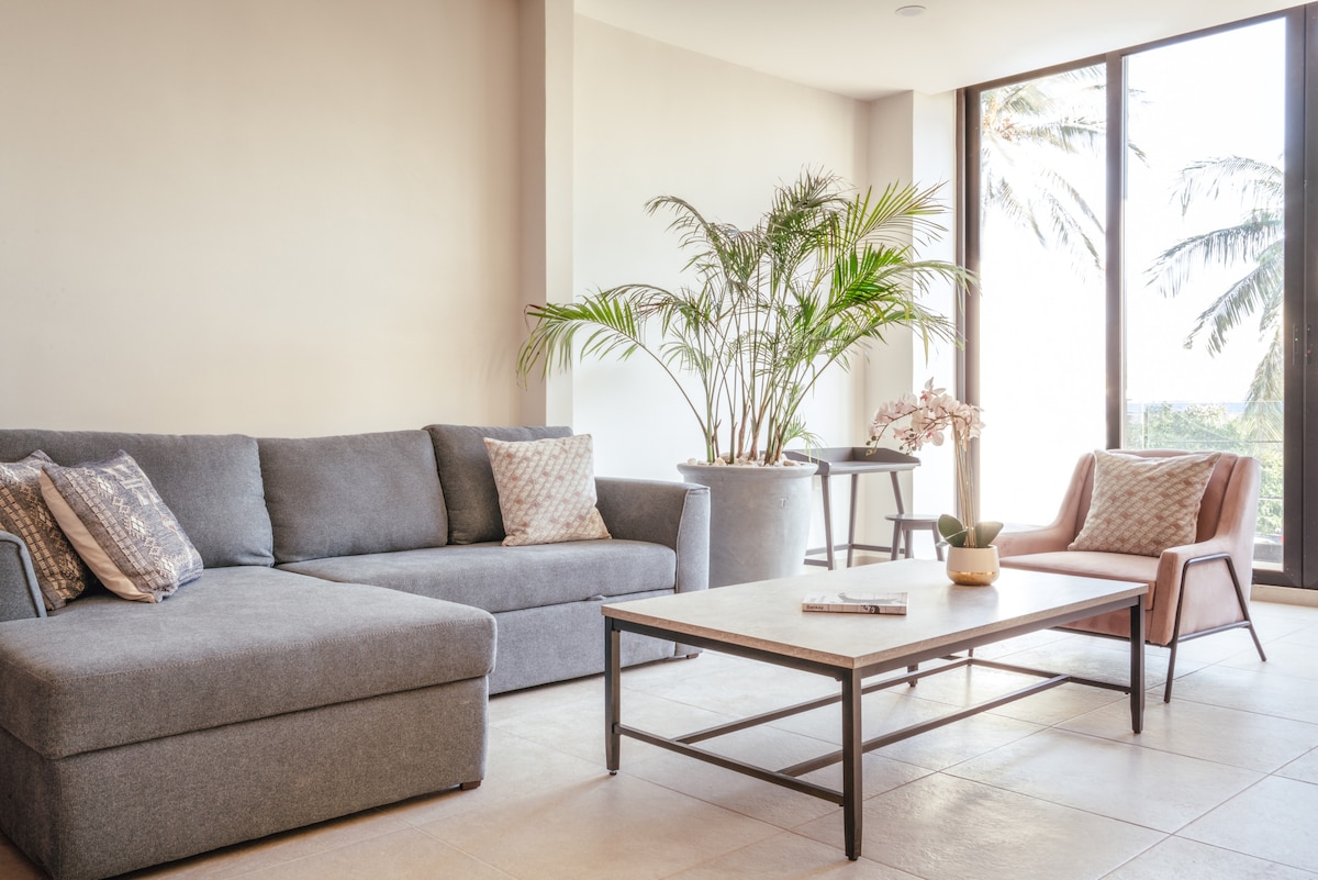 A comfortable living area features a gray sectional sofa with two decorative pillows, a minimalist coffee table, and a light brown accent chair. Large windows allow ample natural light, with greenery visible outside, creating a relaxing atmosphere.