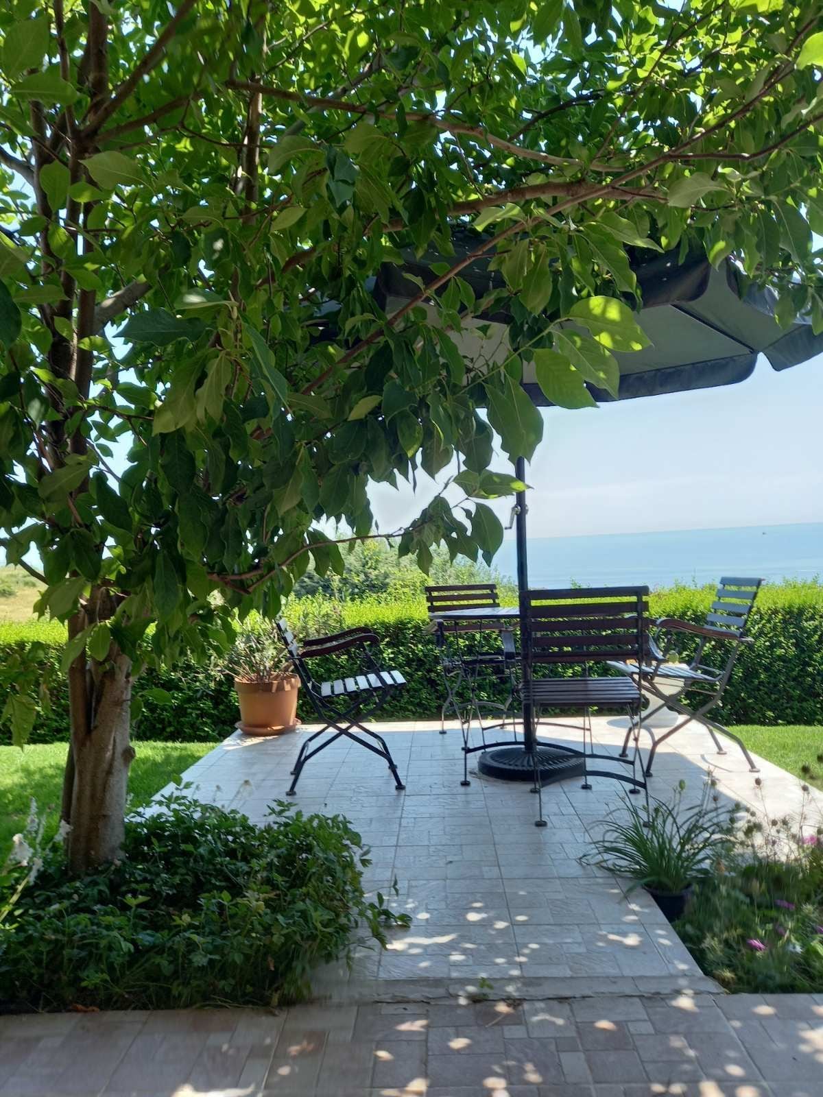 A shaded outdoor seating area is surrounded by lush greenery and colorful plants. Several metal chairs are arranged around a small table, with an umbrella providing protection from the sun. The view of the distant sea enhances the tranquil atmosphere.