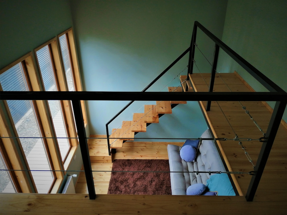 An overhead view of a loft space features a cozy sitting area with a light gray sofa adorned with two blue cushions. A wooden staircase leads down to the lower level, surrounded by large windows that allow natural light to fill the space.