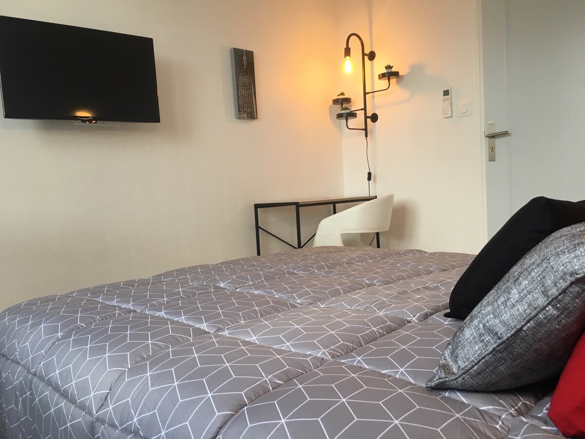 A comfortable sleeping area features a quilted grey bedspread, accented by decorative pillows in black and red. A wall-mounted television is visible, along with a minimalist desk and sleek chair. Warm lighting is provided by a modern wall sconce, creating a cozy ambiance.