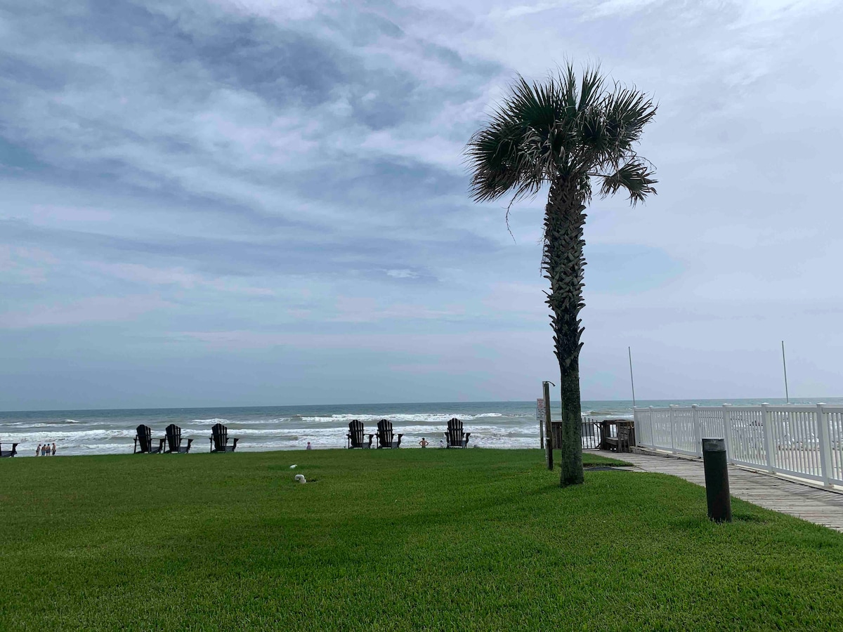 A serene view of the ocean is framed by a palm tree and well-maintained grass. Several wooden chairs are positioned along the shoreline, offering a space to relax. The sky is overcast, adding a tranquil atmosphere to the beachfront scene.