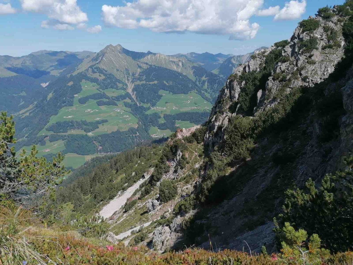 A scenic mountain landscape captures rocky cliffs cascading down to lush green valleys below. The expansive view showcases rolling hills and distant peaks, offering a sense of serenity. Patchy clouds hover above, creating a dynamic sky that enhances the overall depth of the natural panorama.