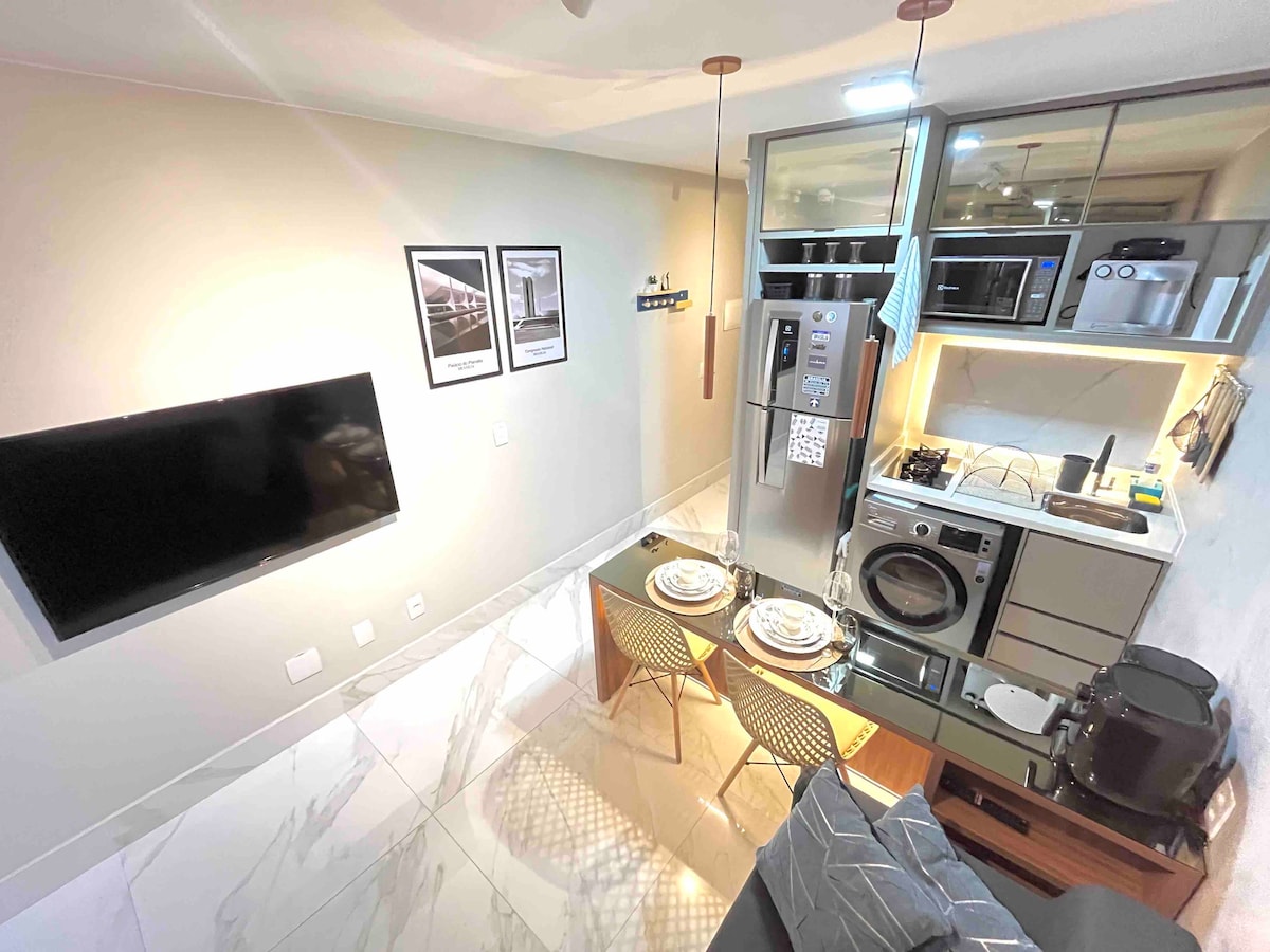 An open living area is presented with a sleek kitchen that includes modern appliances. A dining table is set for two, with dishes arranged neatly. A large flat-screen TV is mounted on the wall, with a cozy sofa nearby, and warm lighting enhances the space.