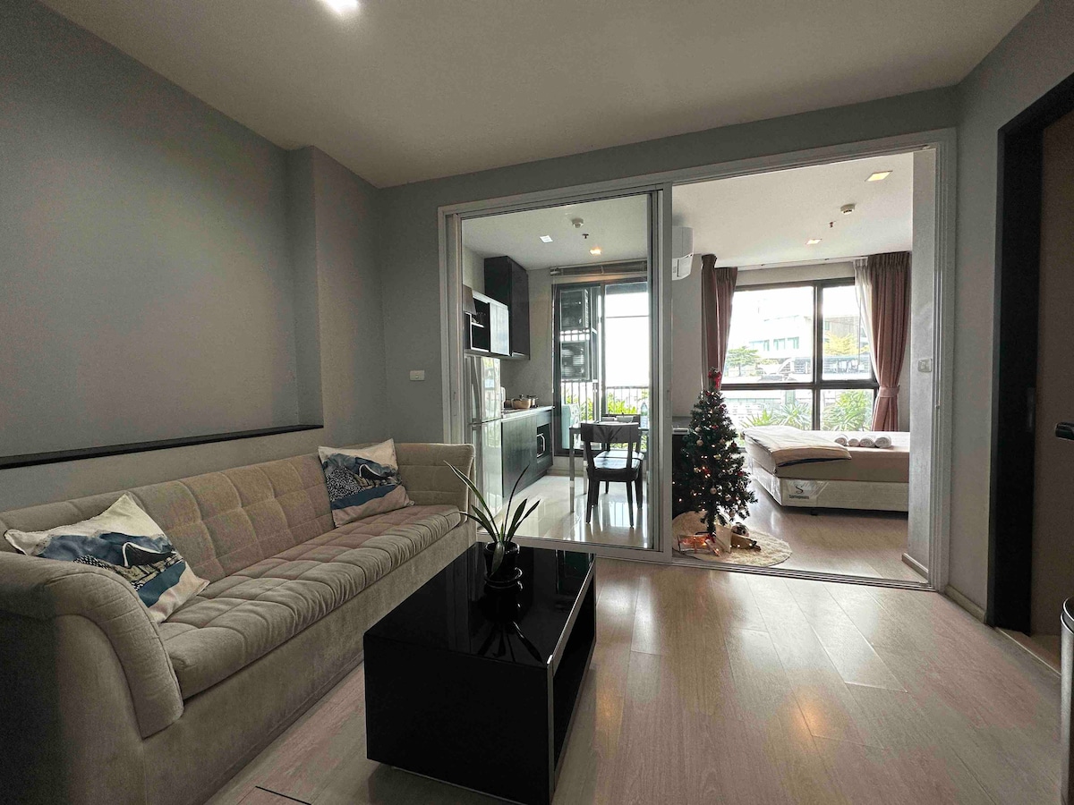A living area features a comfortable sofa with decorative cushions, facing a sleek black coffee table. The space is illuminated by natural light from a large window overlooking a garden, leading to an adjacent dining area. A small holiday tree adds a festive touch.