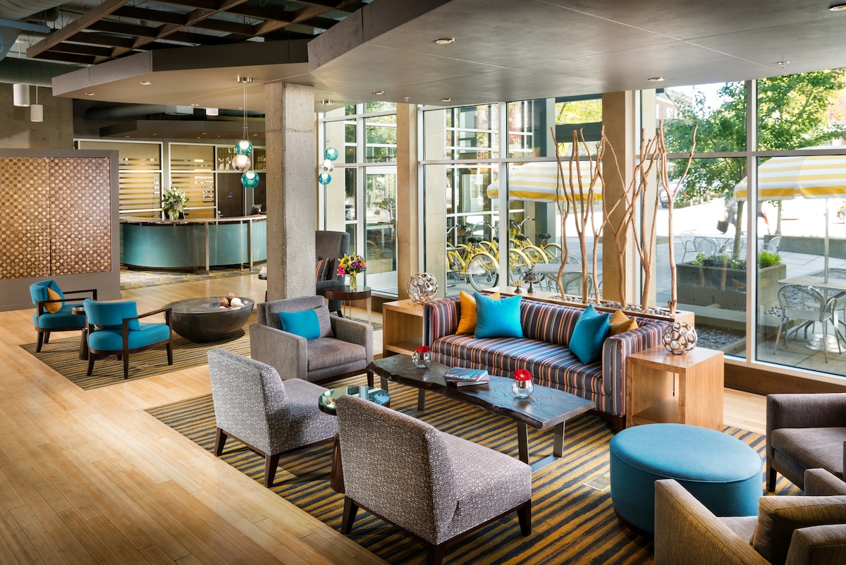 A modern lobby area features large windows allowing natural light to illuminate the space. Comfortable seating arrangements include a striped sofa and various chairs in coordinating colors. Artistic elements like decorative tree installations and table centerpieces enhance the contemporary decor.