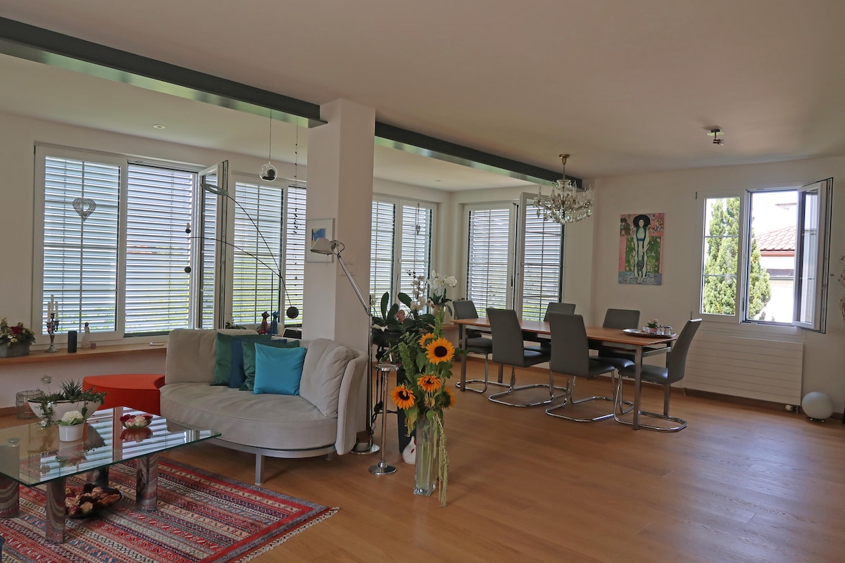A bright living area features a comfortable curved sofa adorned with pillows in a selection of colors. Natural light fills the space through multiple large windows, complemented by a dining table set for six. Fresh sunflowers add a touch of nature, enhancing the overall ambiance.