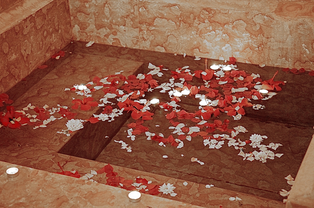 A serene water feature is adorned with floating white flowers and scattered rose petals. Subtle lights illuminate the scene, reflecting gently on the water's surface, while the surrounding stone steps display an earthy texture that complements the tranquil ambiance.