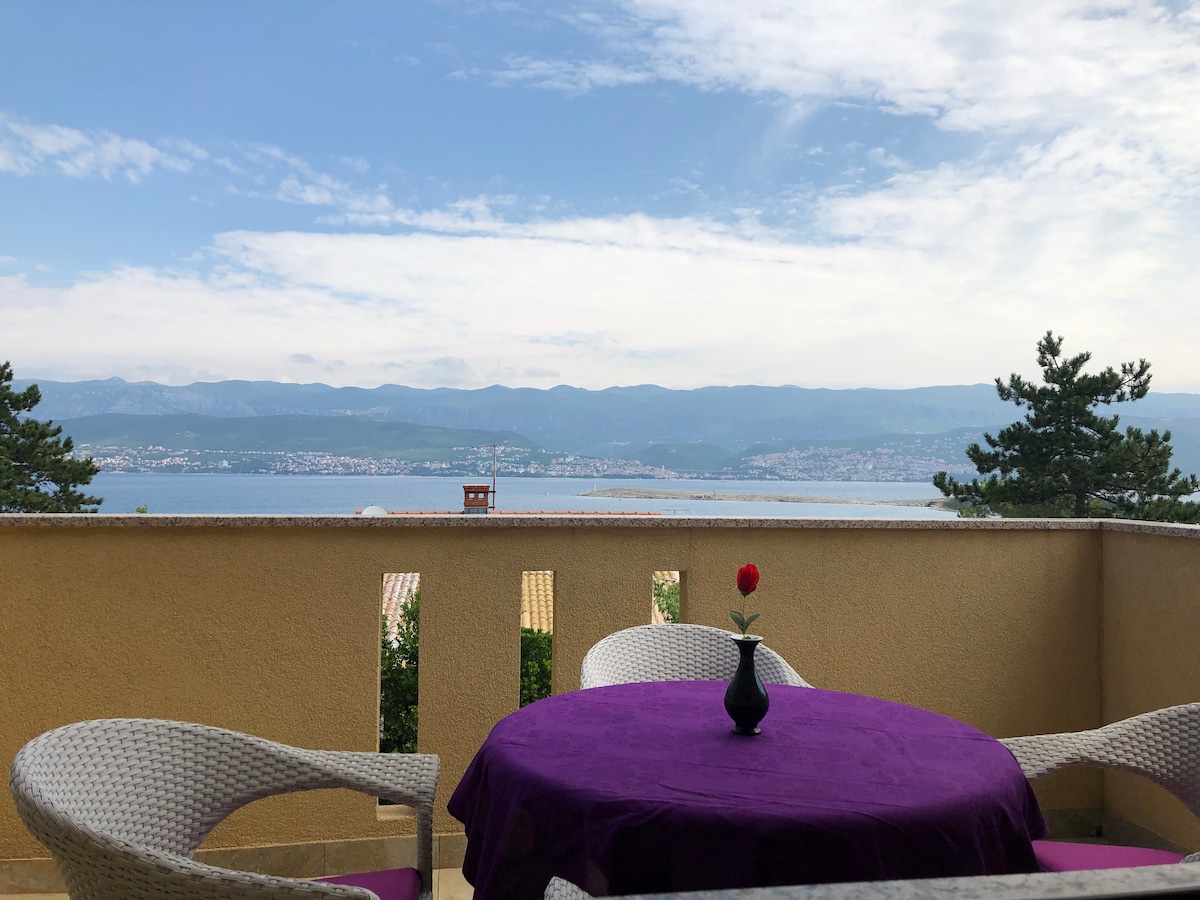 A balcony terrace presents a sea view with a purple table set for leisure. A single flower vase sits centrally on the table, adding a touch of color. The surrounding railing provides a sense of openness, with distant mountains visible under a partly cloudy sky.