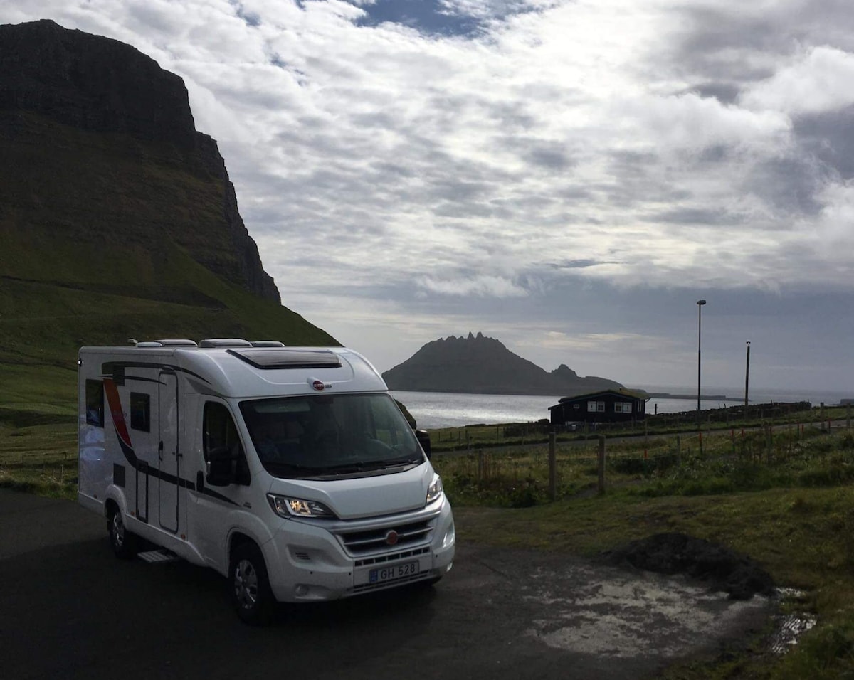 A Bürstner Brevio t646 motorhome is parked on a scenic roadway, with a prominent rock formation rising in the background. The vehicle is positioned against a backdrop of rolling green hills and a cloudy sky, highlighting the natural beauty of the landscape.
