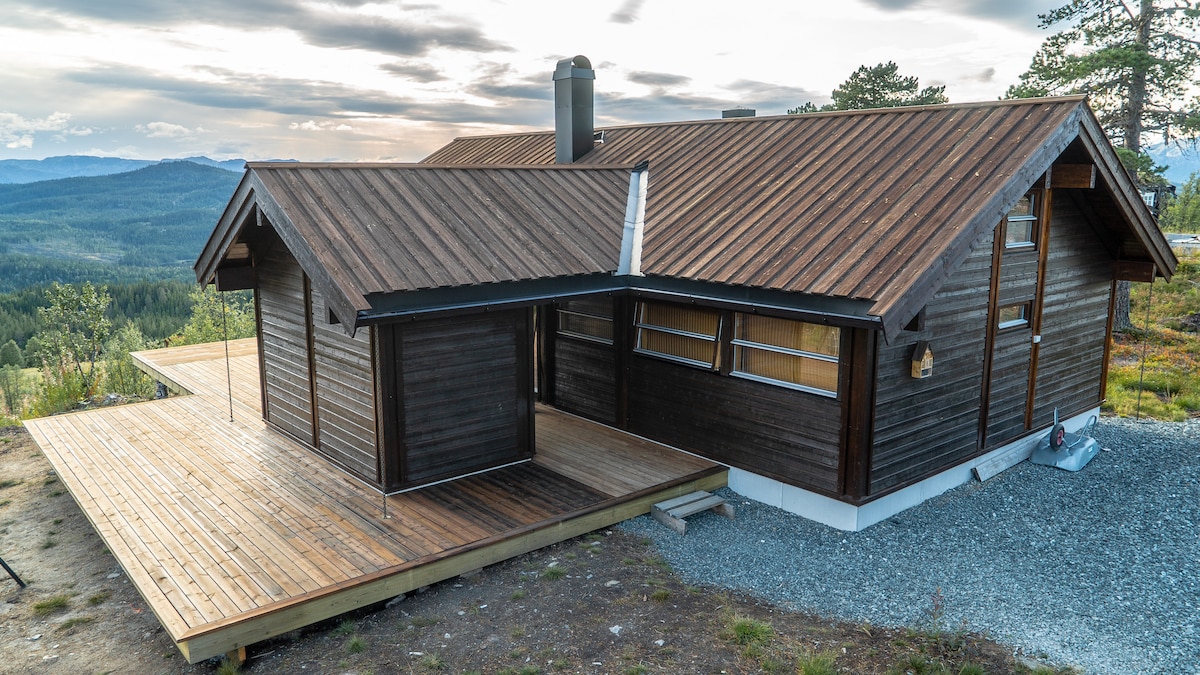 A modern wooden cabin is set on a raised wooden deck, surrounded by a natural landscape. The structure features a sloped roof and large windows that invite ample natural light. A chimney rises from the roof, and gravel pathways lead to the entrance.