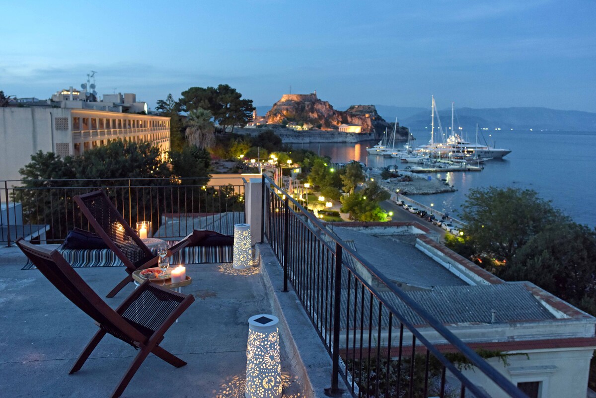 A rooftop terrace offers a serene view of the Ionian Sea and the Venetian fortress at dusk. Two wooden lounge chairs face the water, surrounded by ambient lighting and decorative elements. Boats are visible in the harbor, enhancing the tranquil setting.