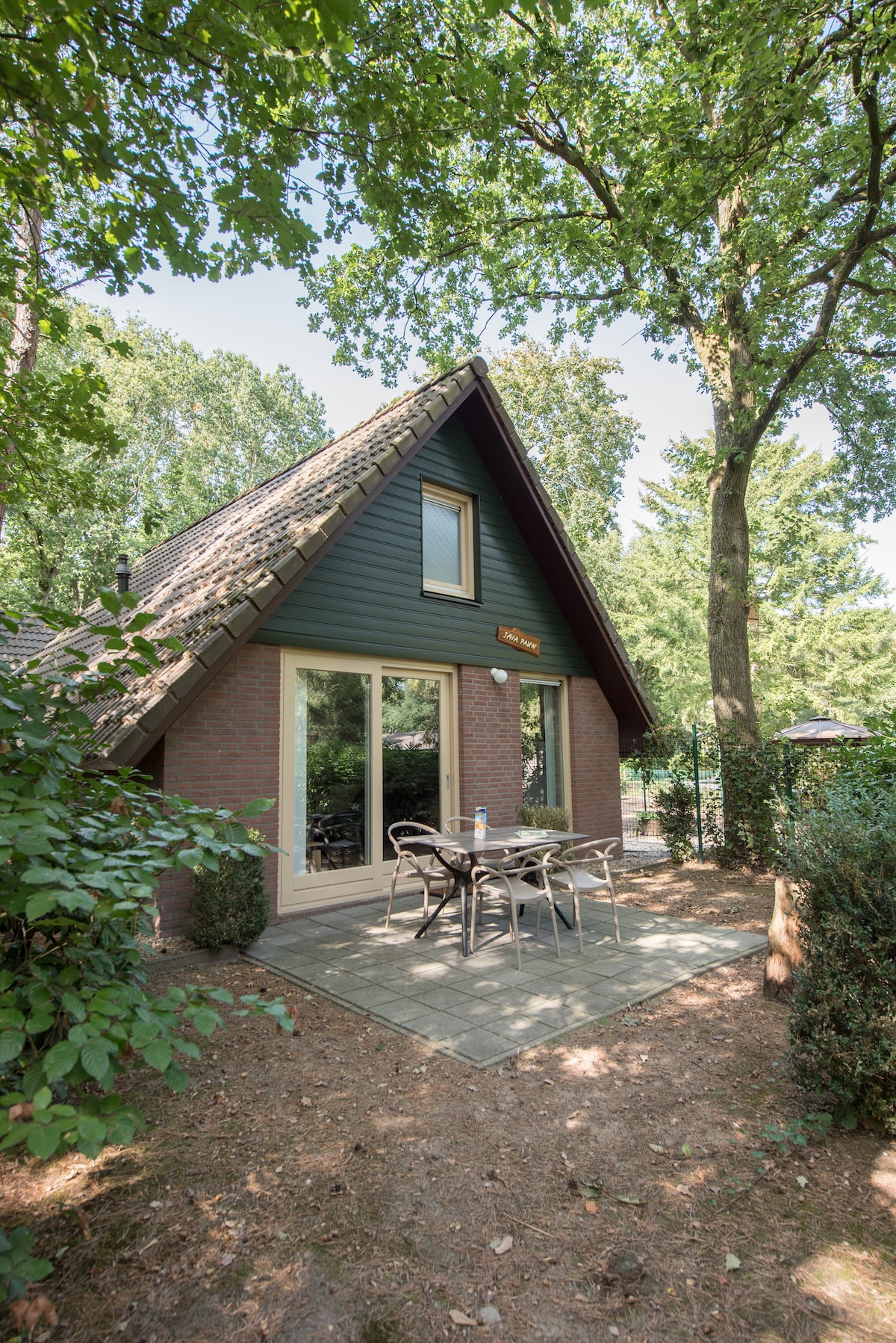 The exterior of the bungalow is surrounded by lush greenery, featuring a sloping roof and large glass doors that open to a patio area. A stone-paved patio hosts a dining table with chairs, providing an inviting space to enjoy the natural surroundings.