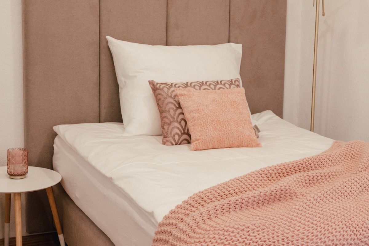 A comfortable bed with a padded headboard is featured in the room. Decorative pillows in soft shades rest on the white bedding. A bedside table with a candle enhances the cozy atmosphere. A warm knitted blanket is draped over the foot of the bed.