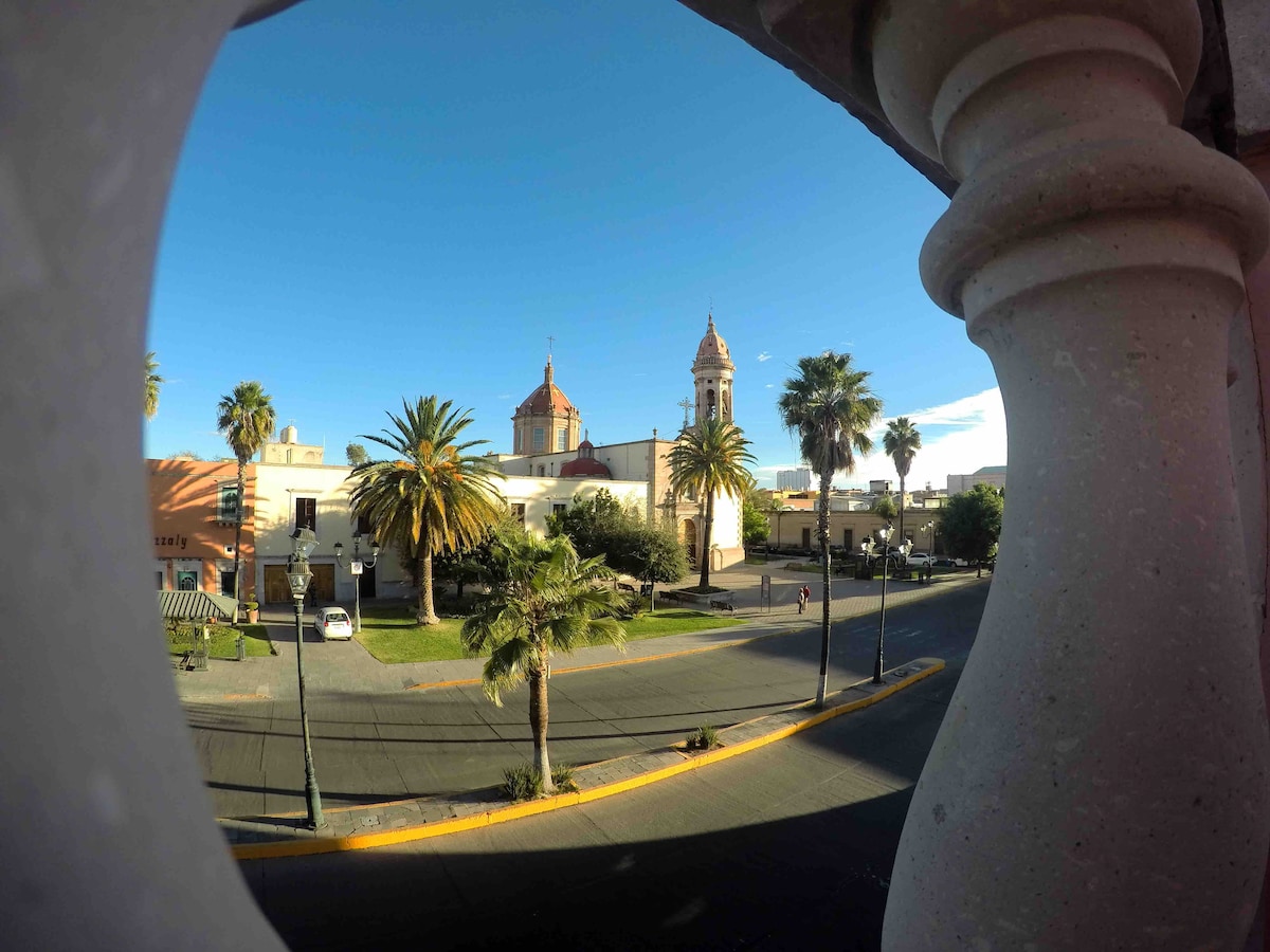 A view of a sunny street adorned with palm trees and colonial buildings is framed by an architectural element. Two church towers with distinctive domes rise in the background, providing a glimpse of the historic surroundings.