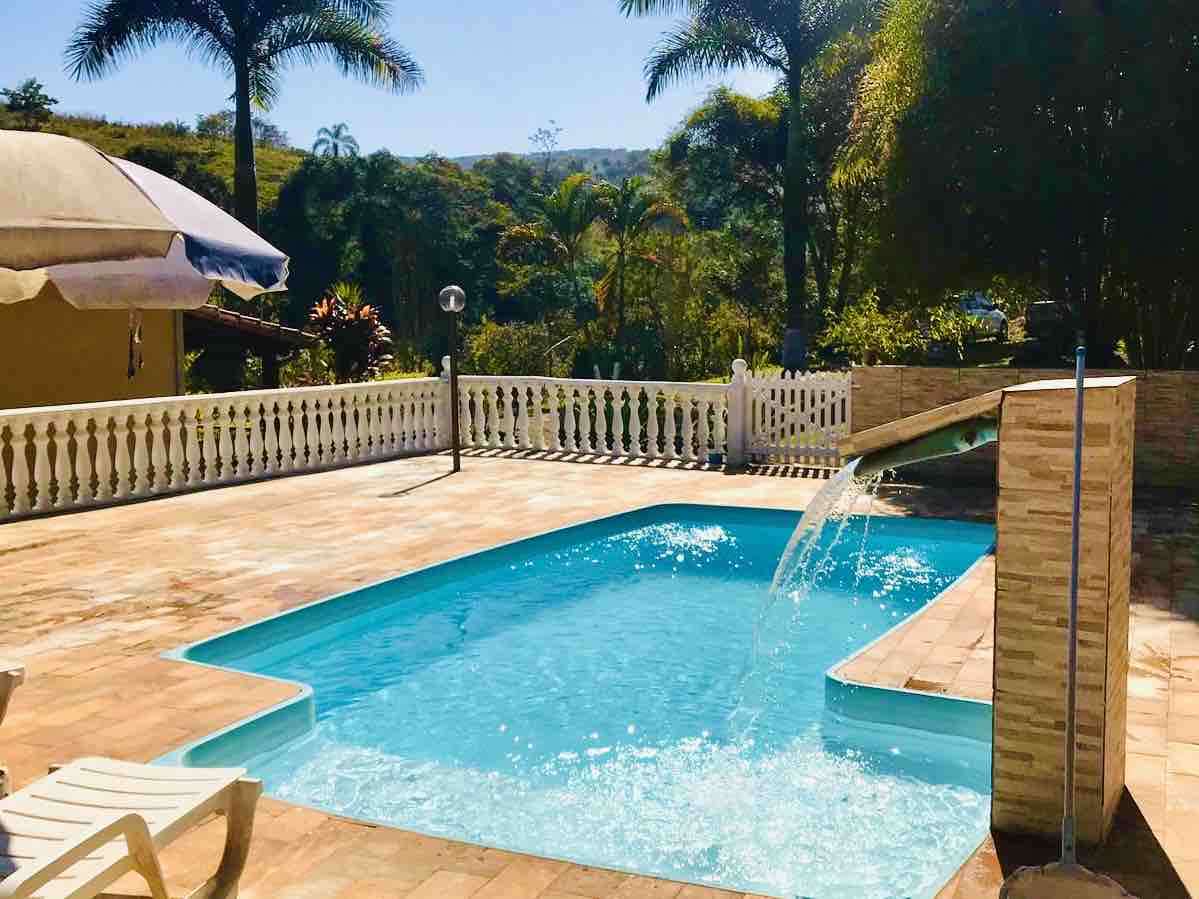 A clear swimming pool is surrounded by a patio with sun loungers and a white fence. A gentle waterfall feature adds a calming sound, with lush greenery visible in the background under a bright blue sky.