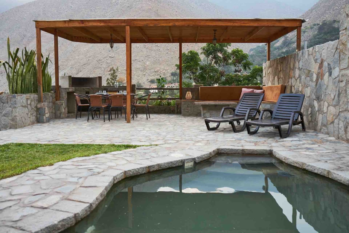 An outdoor seating area is situated under a wooden pergola, with a stone patio and a small pool nearby. Several lounge chairs are arranged for relaxation, while a dining table is set for meals. Lush greenery and mountain views are visible in the background.