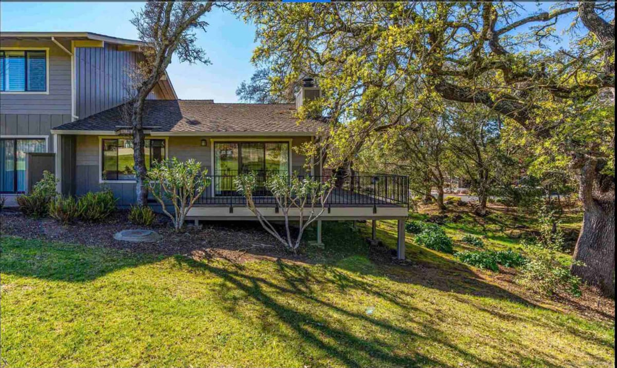 The exterior of the condominium is shown, featuring a wrap-around deck with railings and a landscaped yard. The structure is framed by trees, providing shade and a natural setting, while ample sunlight highlights the greenery and creates shadow patterns on the ground.