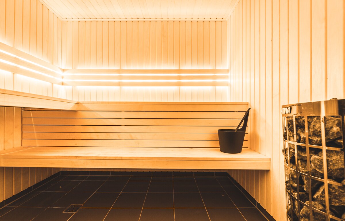 A wooden sauna space features light-colored slatted walls and benches. A bucket is placed on one of the benches, while a metal stove stands in the corner, filled with stones. Subtle lighting enhances the warm ambiance within the enclosed area.