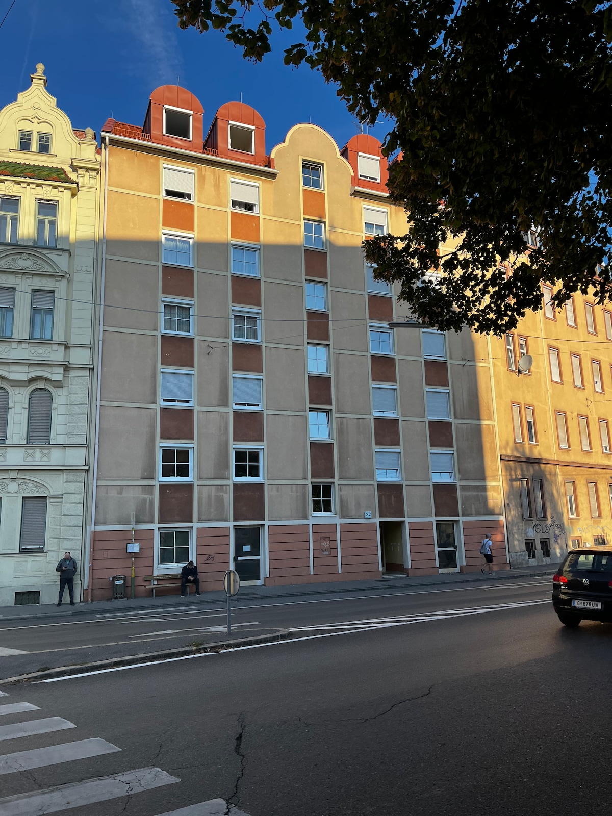 A multi-story building presents a blend of modern and classic architectural styles. The facade features a combination of muted earth tones, large windows, and a unique rooftop line. Sunlight creates shadows, enhancing the structural details against the clear blue sky.