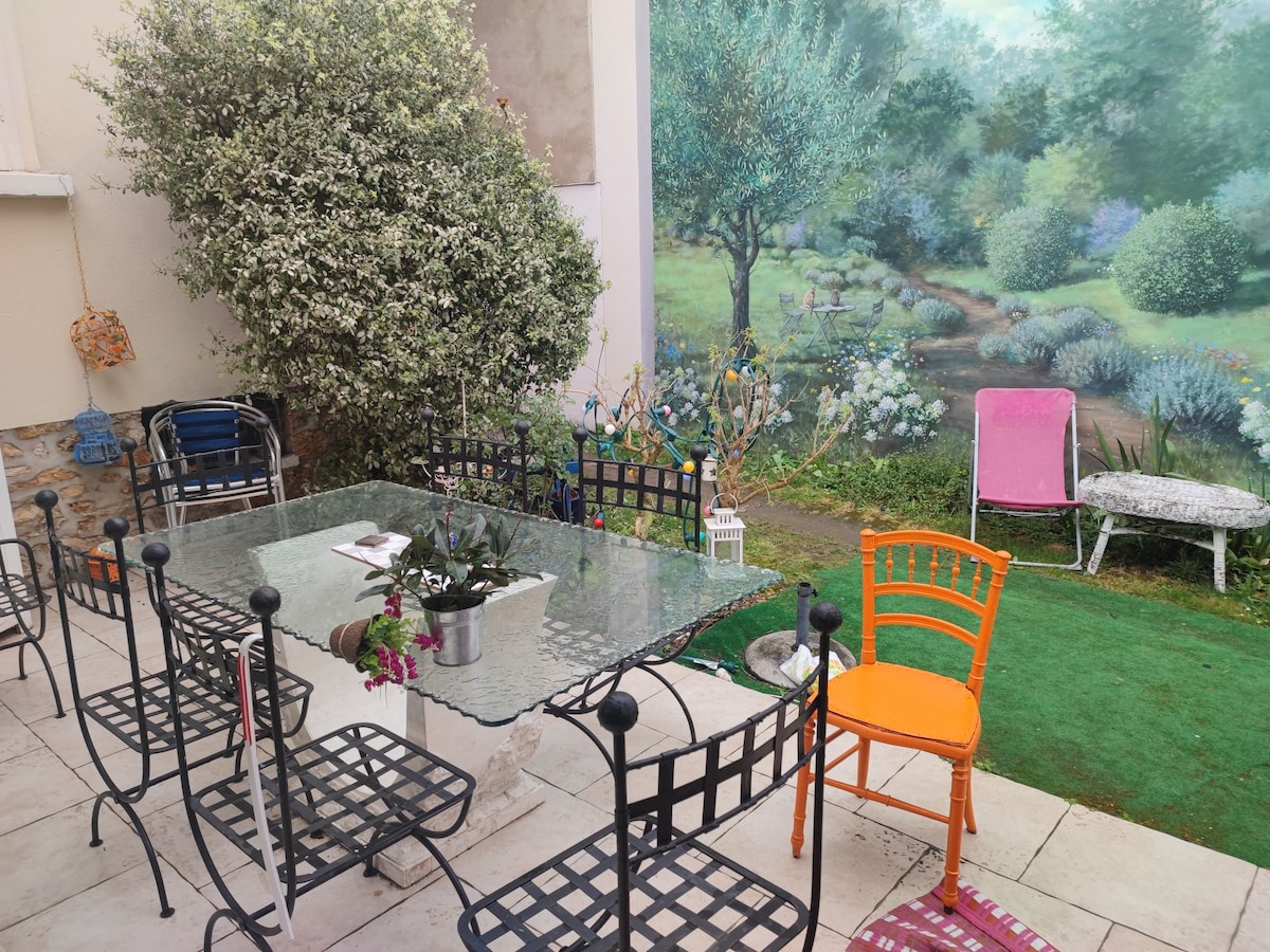An outdoor area features a glass-topped dining table surrounded by black metal chairs. Lush greenery and a vibrant mural of a landscaped garden enhance the space, while one orange chair adds a pop of color. Potted plants and garden decorations are also visible.