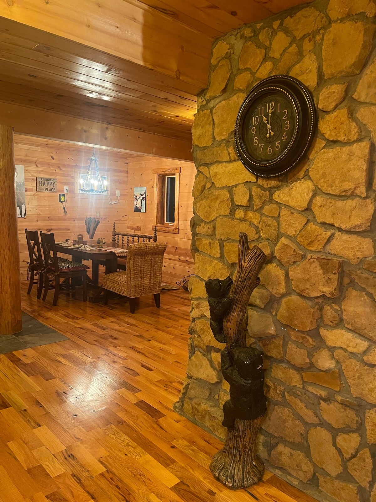 An inviting entryway features warm wooden paneling and a stone accent wall with a clock. A dining area is visible, furnished with a wooden table and chairs, adjacent to a cozy seating space. Two bear sculptures climb a tree trunk, adding rustic charm.